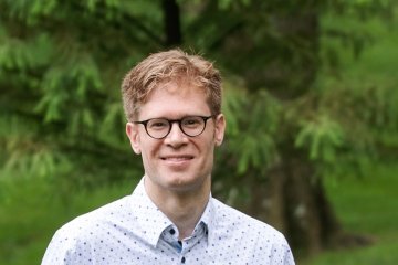 Man with dark blonde hair with glasses and light blue dress shirt in front of tree