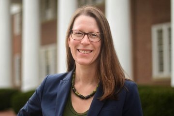 Lady with dark brown shoulder length hair with glasses, wearing navy blazer and dark top in front of Old Centre