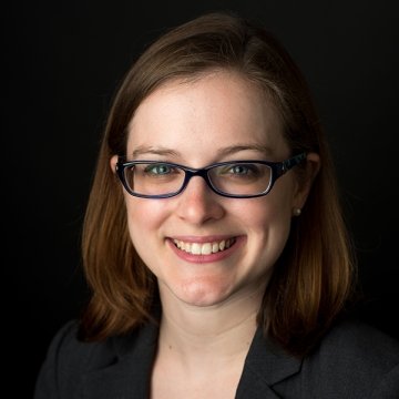 Lady with short brown hair wearing glasses and black jacket standing in front of dark background