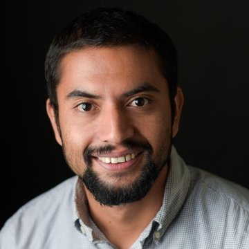 man with dark hair and gotee wearing grey button up top in front of black background