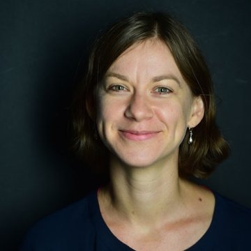 Lady with short dark hair wearing earrings and black top in front of black studio background