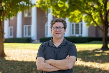 Man with curly dark hair wearing glasses and grey polo with arms crossed standing in front of old centre