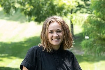 Lady with shoulder length brown hair wearing black short sleeve top standing outside on campus