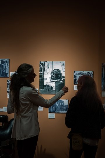 Seen from behind, two people look closely at black-and-white photos displayed on an orange wall. 