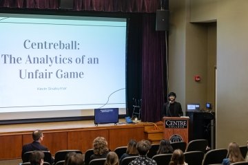 A student stands at a podium in front of a seated crowd. On a screen behind him are the words "Centreball: The analytics of an unfair game."