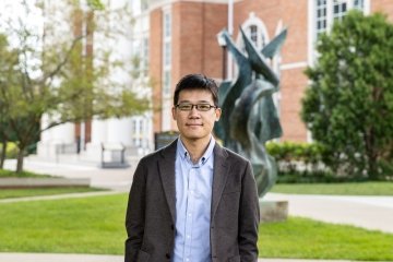 Man with dark hair wearing glasses, sports coat and button up shirt in front of the flame statue