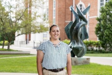 Lady with pulled back pony tail wearing striped top with khaki pants in front of the flame statue