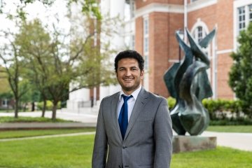 Man with dark hair wearing grey suit jacket and white shirt with navy tie in front of the flame statue