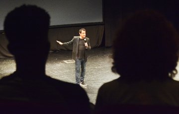 A man holding a microphone is seen between the silhouettes of two audience members. 