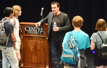 Students carrying backpacks stand in a loose circle around a man holding a water bottle and resting his arm on a podium bearing the Centre College logo.