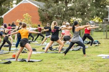 Students are seen from behind as they hold the same yoga class on a green lawn with trees and a brick building in the background.