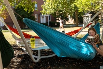 A student reads a book while lying in a blue hammock suspended in the shade with brick buildings in the background. 