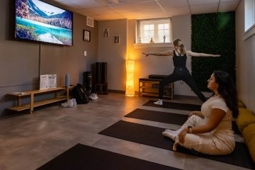 Two students do yoga poses on mats in a room equipped with a large TV and yoga equipment. 