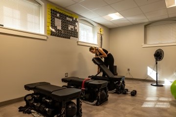 A student adjusts one of three workout benches in a small room containing exercise equipment. 