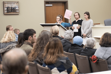 A professor holds papers aloft as she and students stand behind a lecturn in front of a large group of seated people.