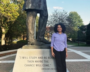 Lady with dark hair wearing button up dress shirt and dark pants standing next to Lincoln statue