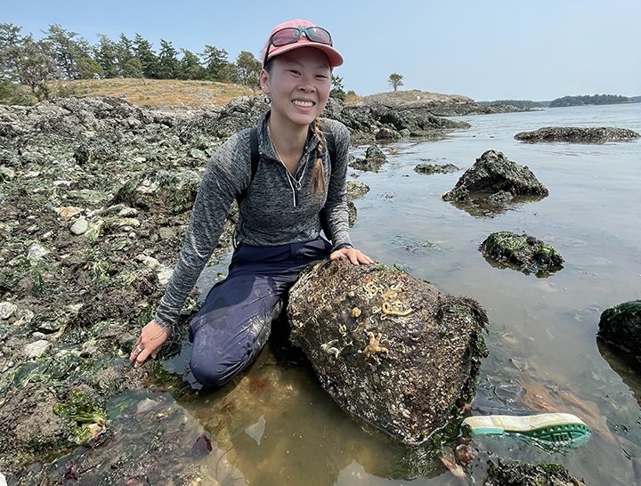 A young woman kneels behind a large stone where a rocky beach meets the ocean. 