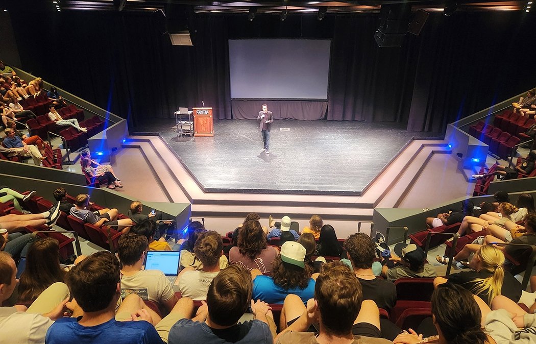 A man talks into a microphone while standing on a stage surrounded by theatre seats filled with students.