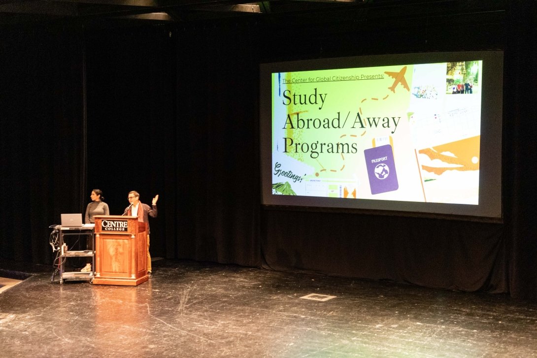Two people stand behind a podium while a projection screen introduces Centre College Study Abroad & Away programs. 