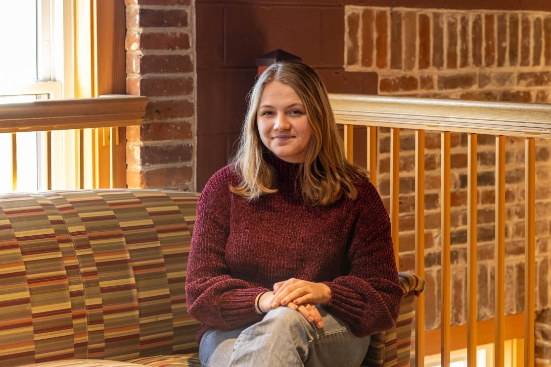 Alli Barrows '25 - student sitting on couch wearing maroon sweater and jeans with long brown hair