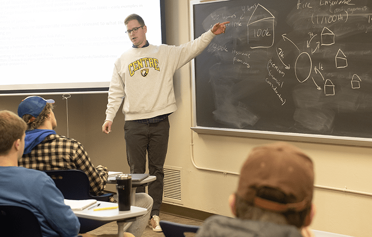 Derek Nafziger wears a Centre College sweatshirt as she stands at a black chalkboard teaching students about real estate insurance. 