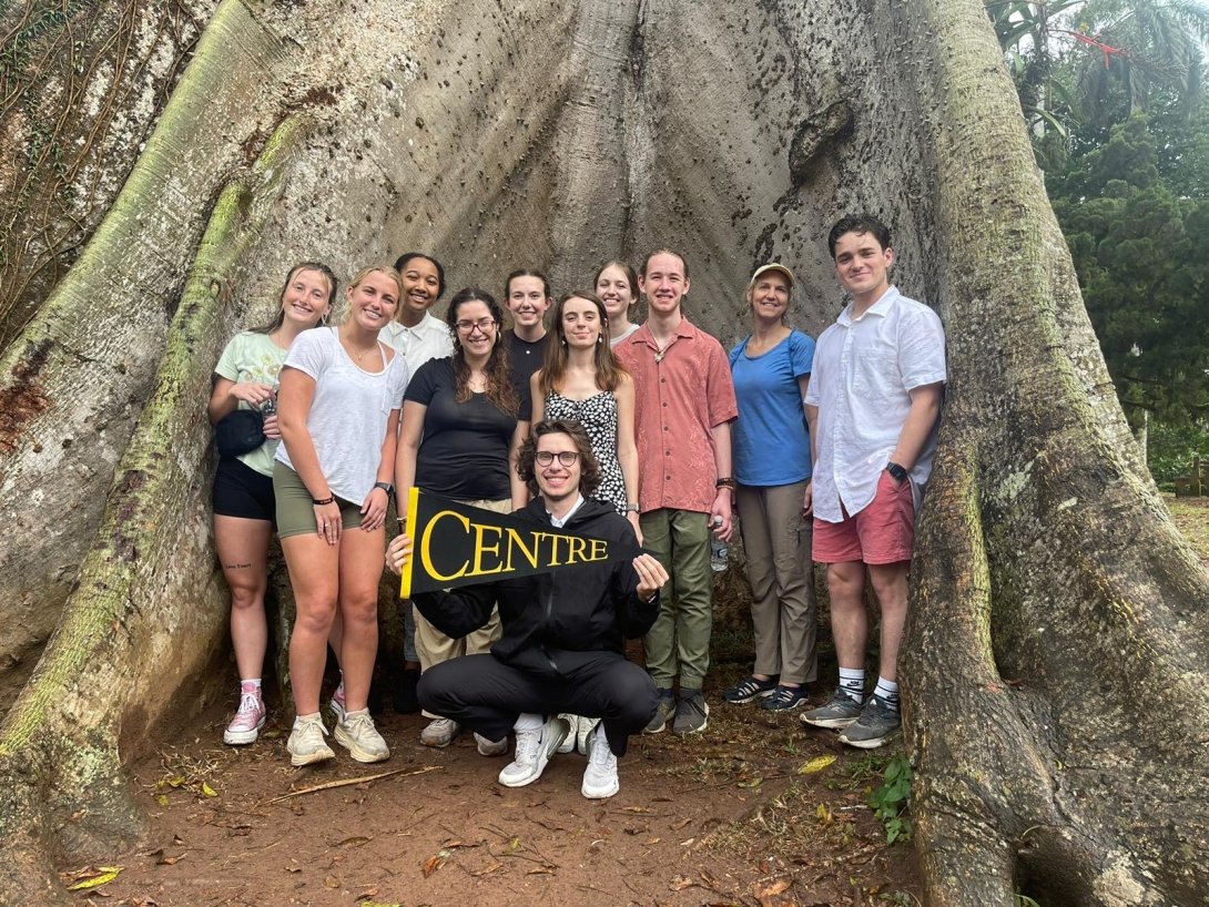 CentreTerm students in Ghana - group of students standing in front of large tree roots. One student holding Centre pennant.