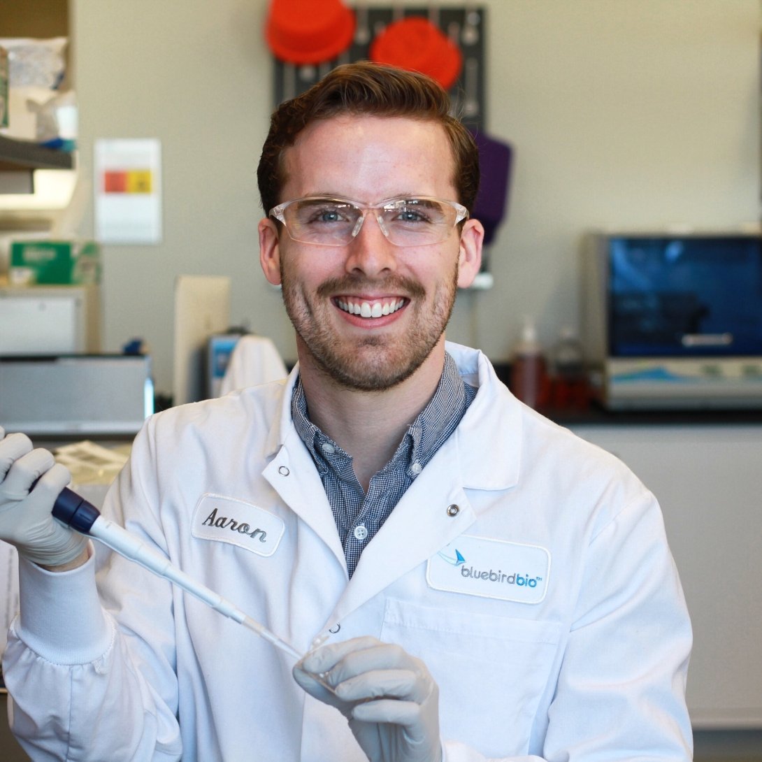A man wearing a white lab coat holding scientific instruments smiles for a photo. 