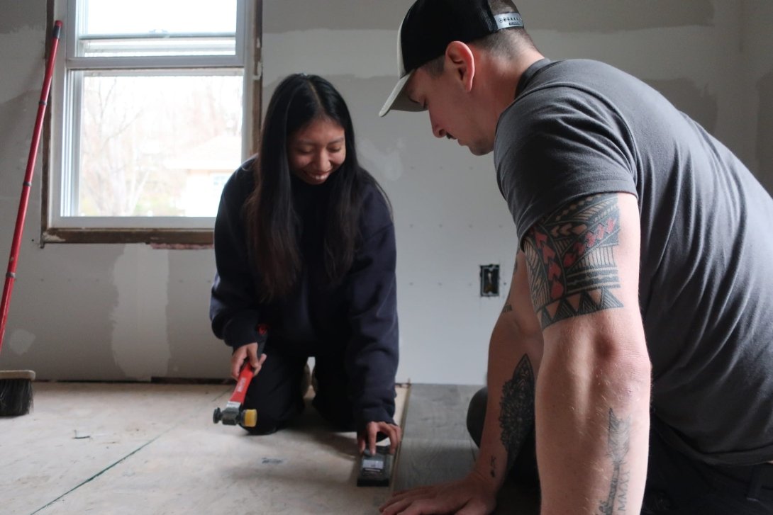A student and staff member are working together on a flooring project in North Carolina. The student is kneeling, holding a hammer and flooring tool, while the staff member leans forward to assist.