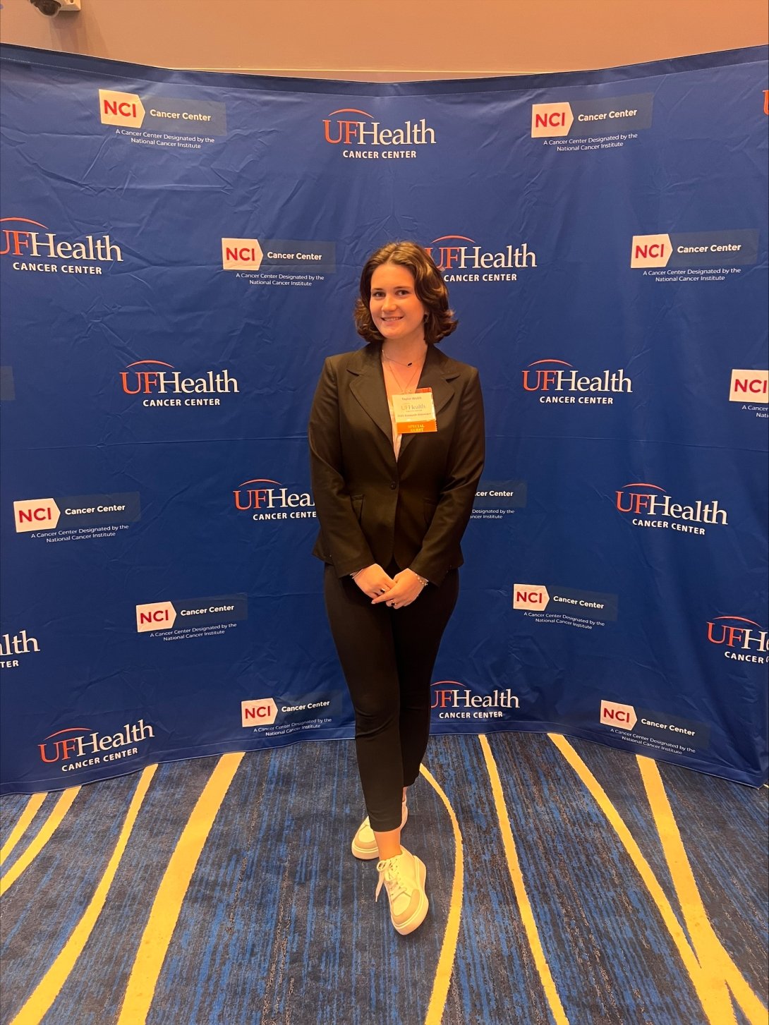 A young woman in a dark business suit poses in front of a conference backdrop with "UF Health Cancer Center" written on it. 