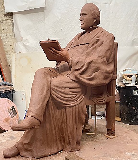A clay sculpture model shows a man in judge's robes seated while writing on a tablet.