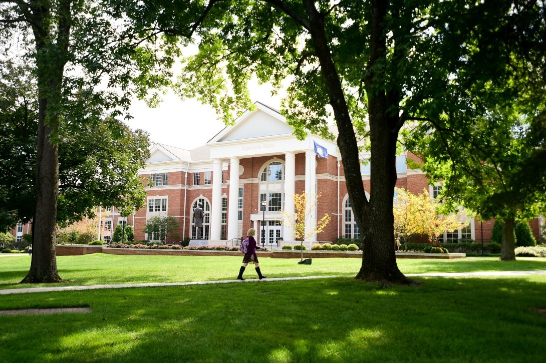 A student walks along a path lined with trees. In the background is Crounse Hall, a large brick building with tall columns.