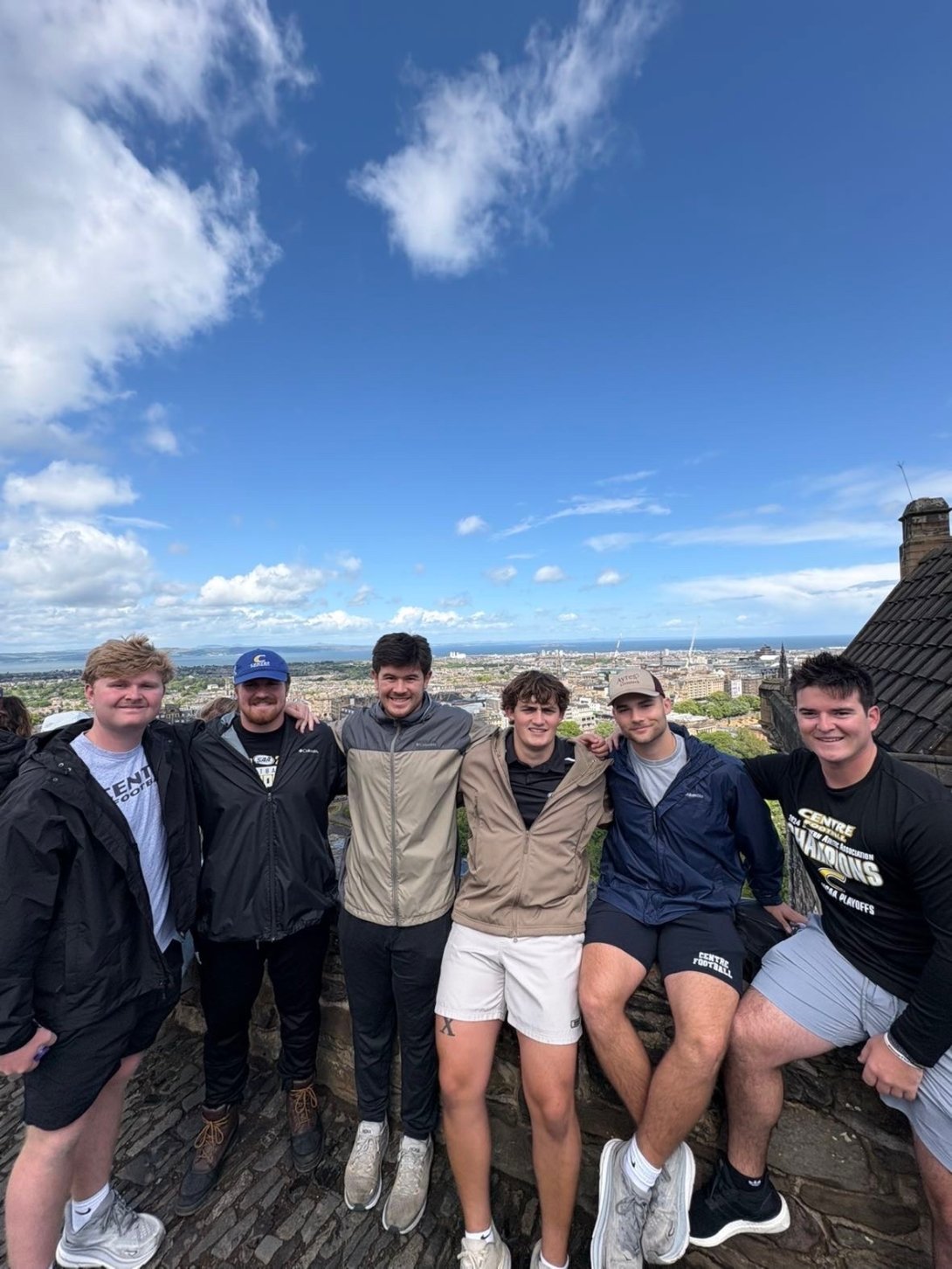 A group of 6 men smile for a photo. They are on a roof of a building with a view of a village behind them.