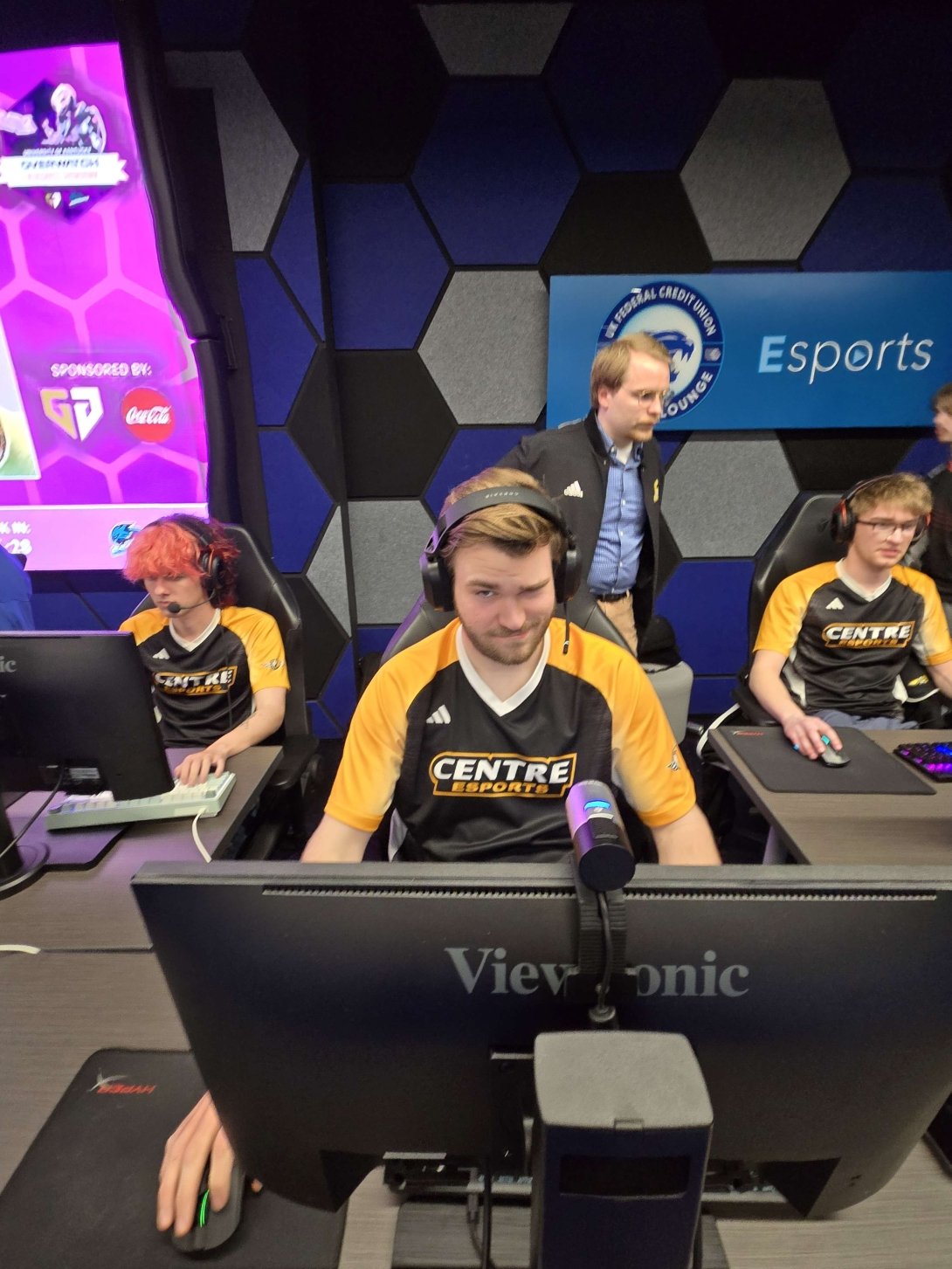 A man in a black and gold Centre College jersey if flanked by two teammates as he competes in an Esports tournament. 
