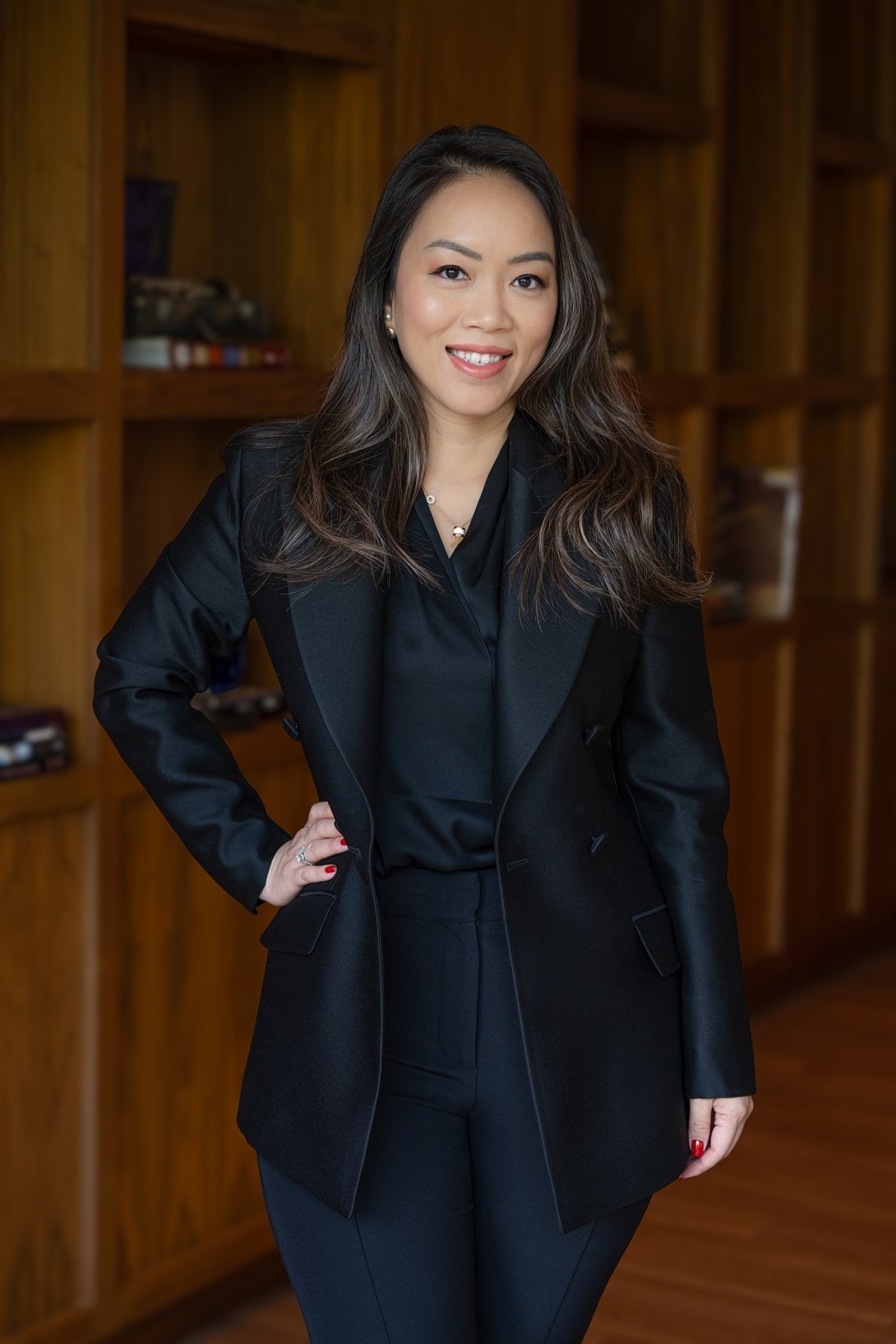A woman ina. dark blazer and black button-up shirt poses for a photo in front of a wall of wooden shelving 