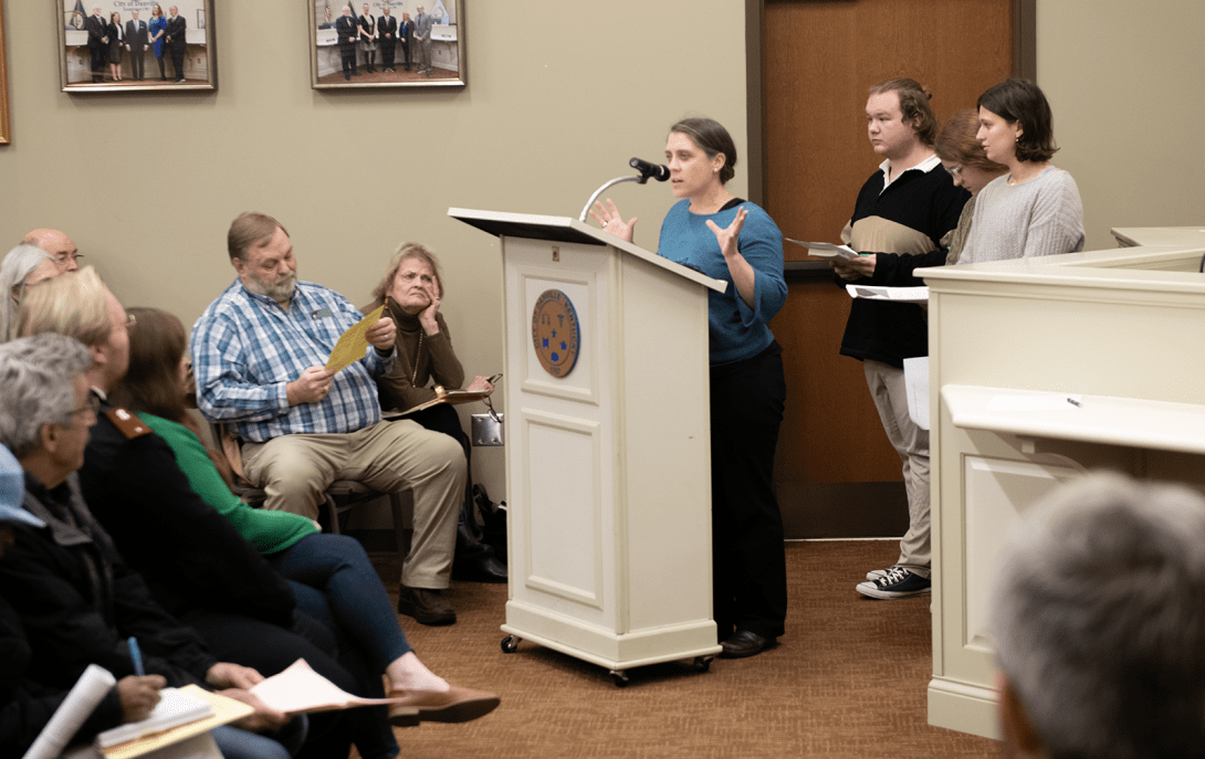 A woman speaks into a microphone at a lectern in Danville City Hall as students stand behind her and a seated audience looks on. 