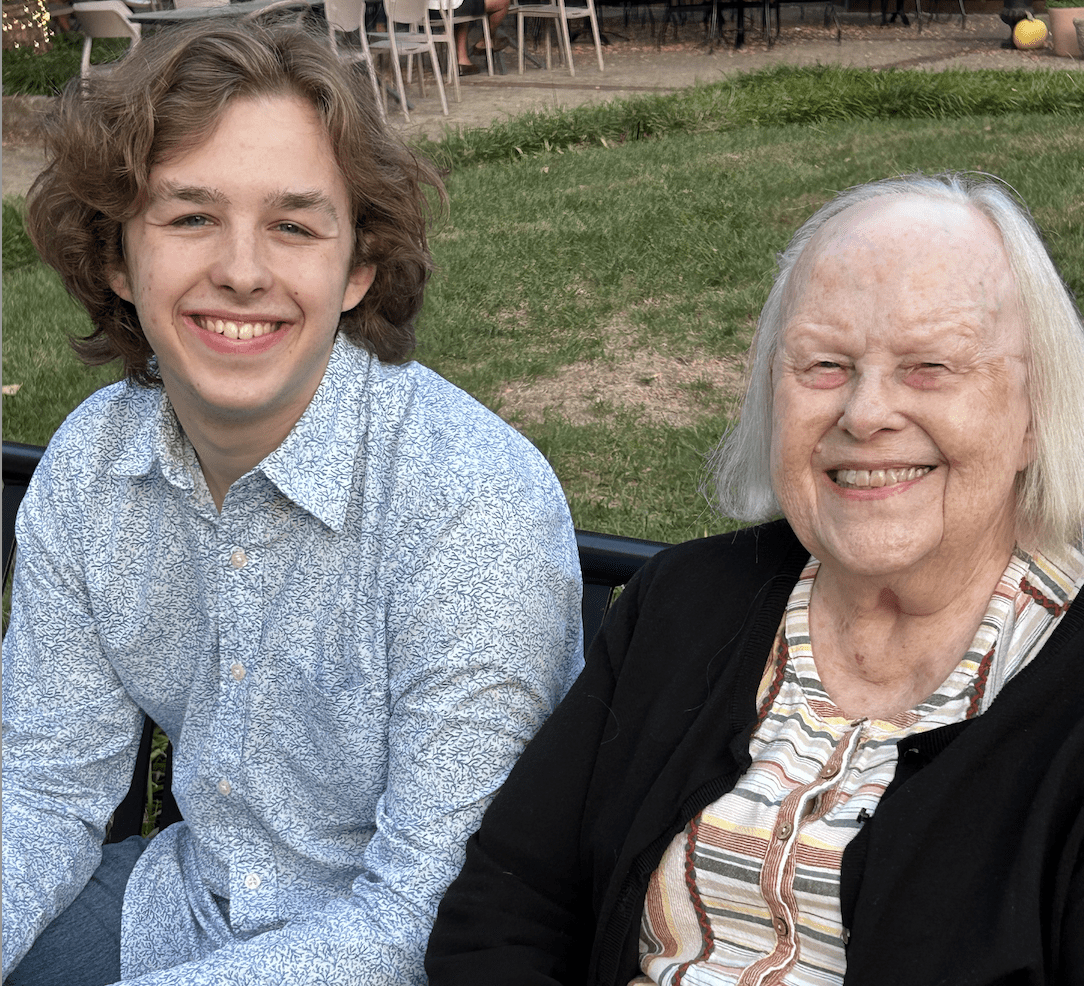 A college-aged man in a blue shirt sits outside next to his grandmother as both smile for the photographer. 