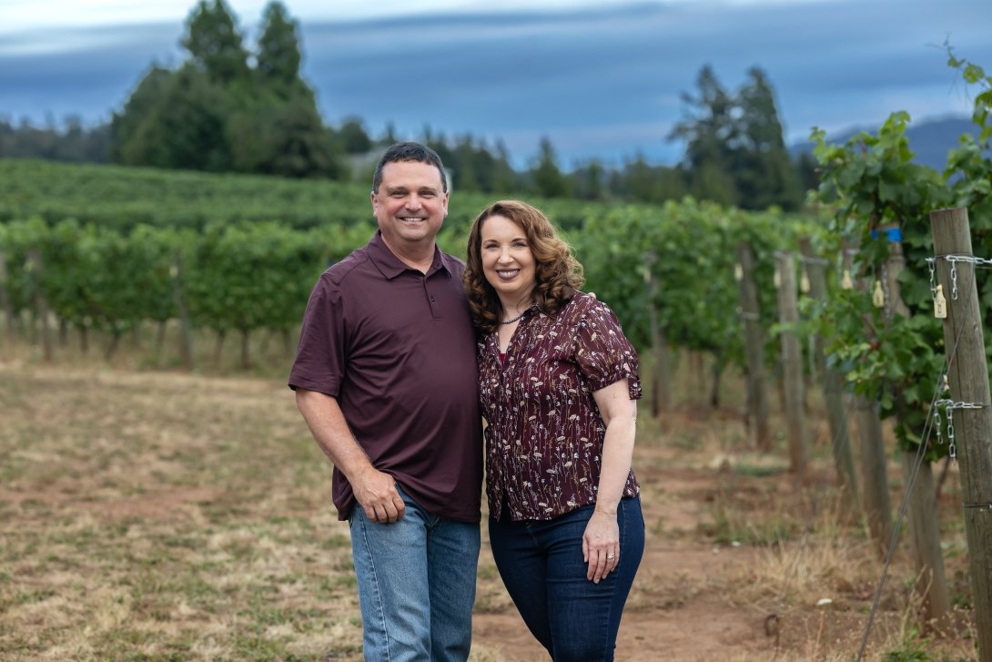 A man and woman post side-by-side in front of a field of wine grapes. 
