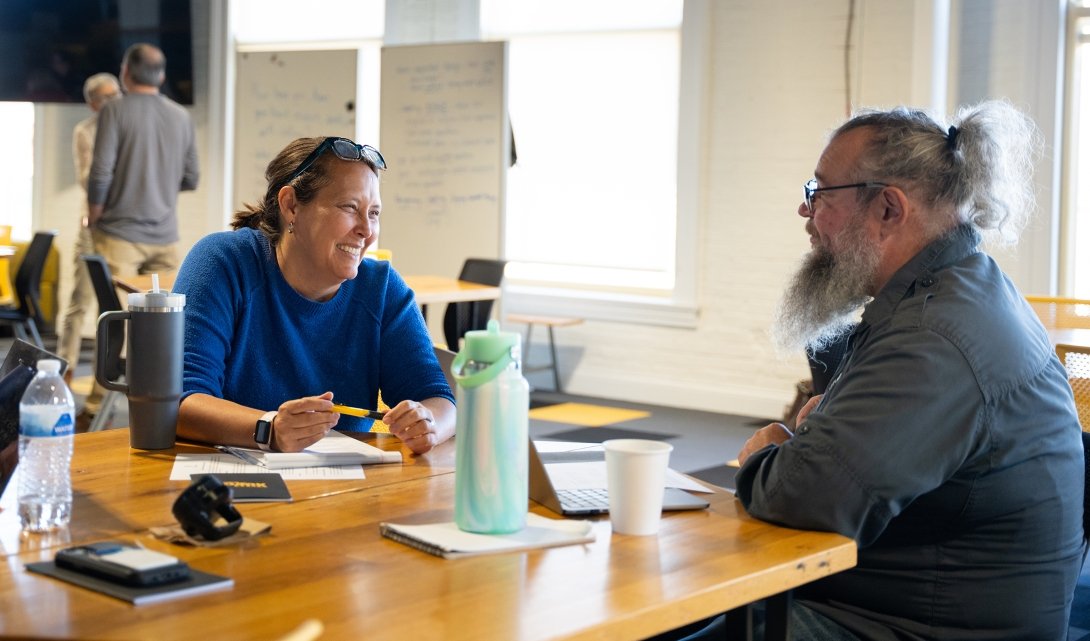 A woman smiles as she speaks to a man while both are seating at a conference table. A man and woman speak together in the background. 