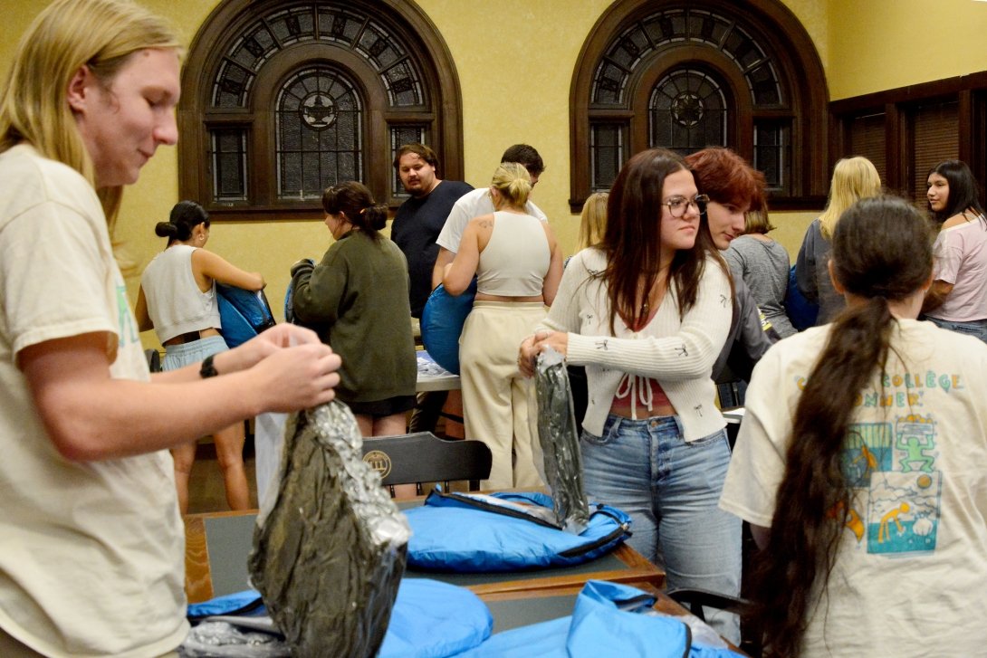 A group of students are standing at tables, packing emergency kits for unhoused individuals.