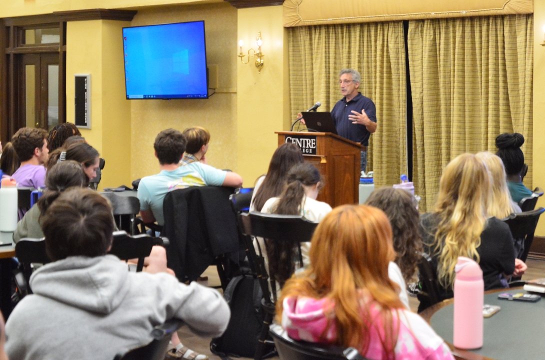 A professor is sitting at the front of the room, presenting to a group of students.