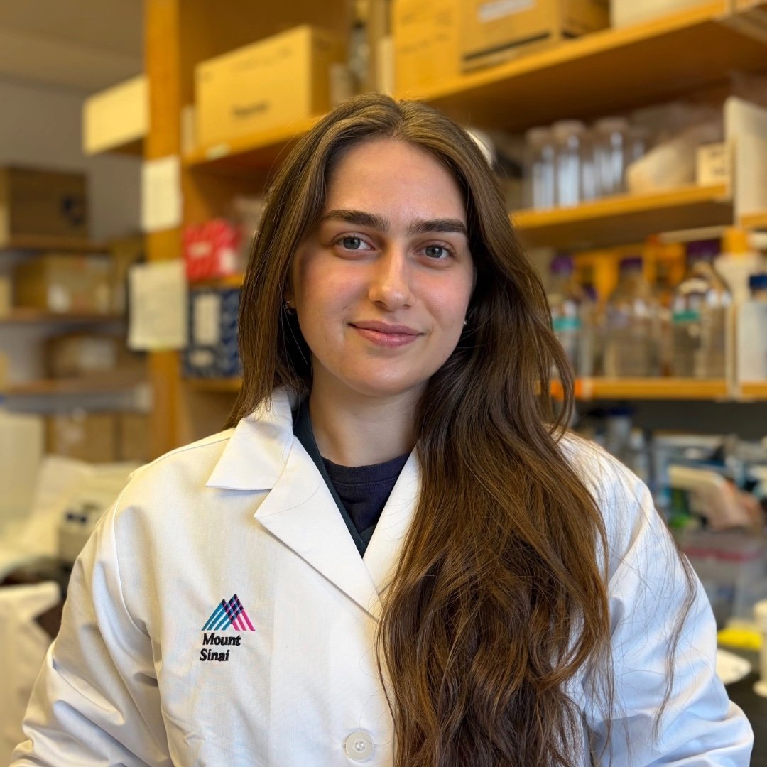 Anna Bostoganashvili is smiling and posing in a lab. Behind her is a shelf of research equipment. She is wearing a lab coat that says Mount Sinai.