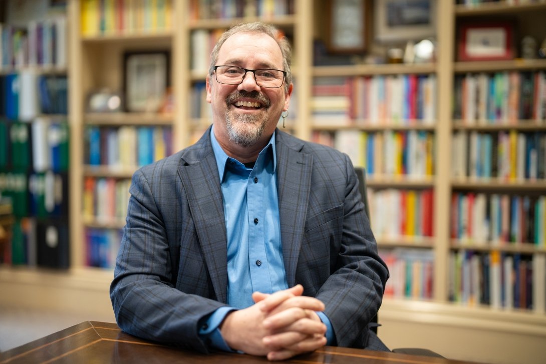 Centre College Provost and Vice President of Academic Affairs Alex McAllister sits at a table with a large bookshelf in the background. 