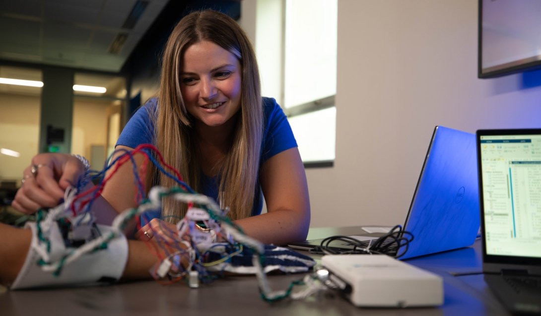 Madison Bates adjusts a device with man wires attached to it that is strapped to the hand of a person off camera as a computer screen shows data.